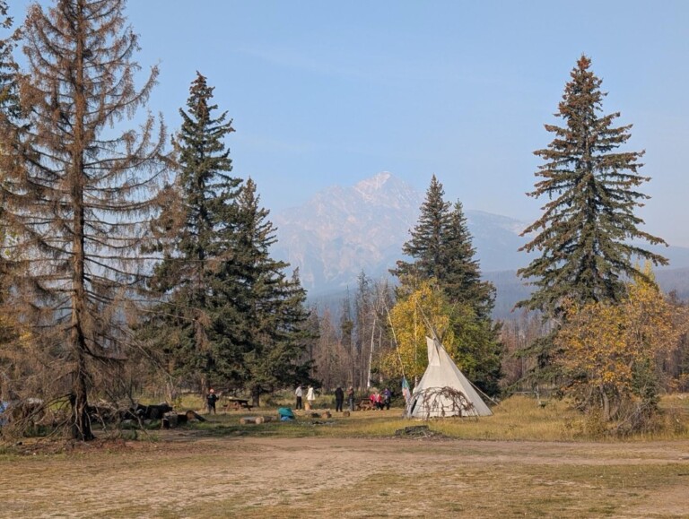 The Jasper National Park’s Cultural Use Area is shown with a group of people next to a tipi surrounded by tall trees. A large mountain is seen in the background through hazy skies.